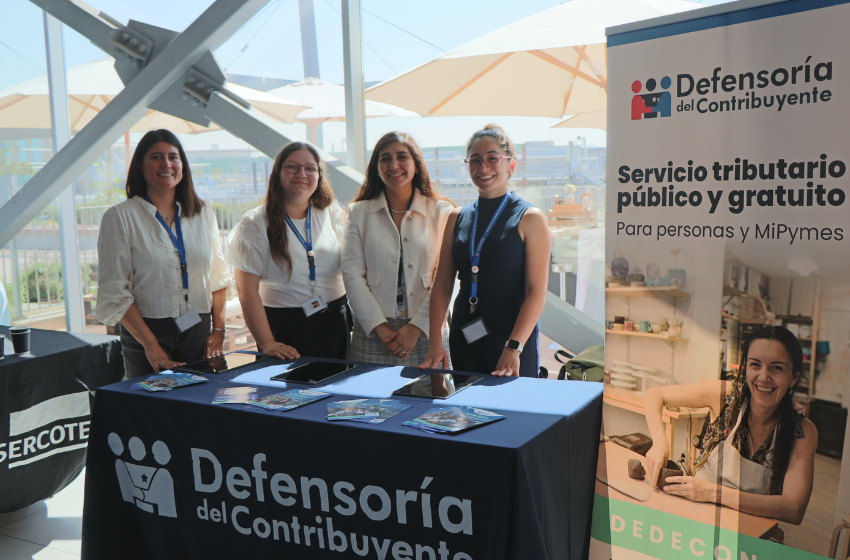 Four women stand behind a Defensoría del Contribuyente information desk at a booth, smiling for the camera in a bright venue.