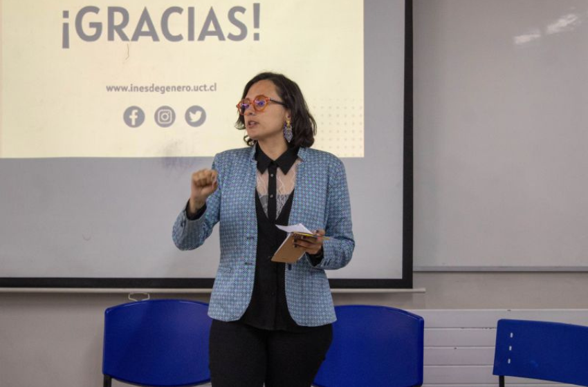 Presenter gives a talk in a classroom, standing by a screen that reads ¡GRACIAS!, holding notes in one hand and gesturing with the other.